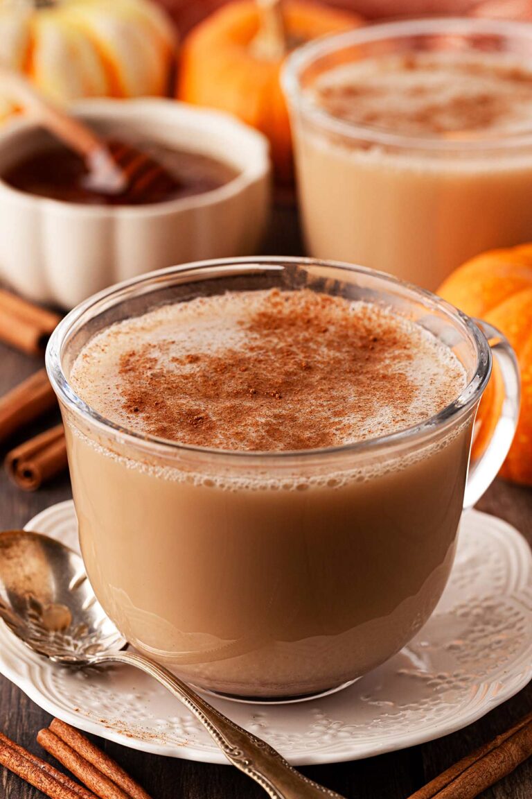 Front focus. A glass mug filled with Pumpkin Spice Chai Latte sits on a saucer with a spoon at it's side.