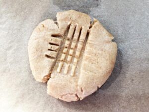 A flatted peanut butter cookie laying on a parchment-lined cookie sheet. It's marked with fork marks.