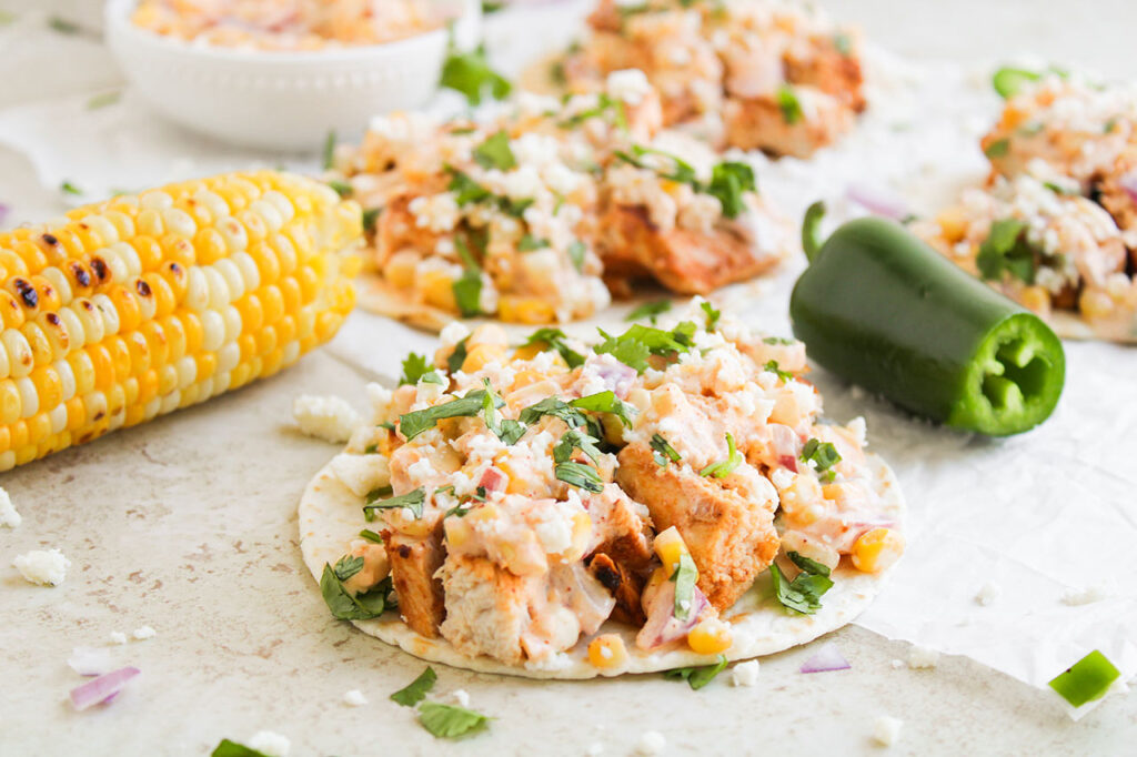 Front focus. Several Street Corn Chicken Tacos lined up on a white surface.