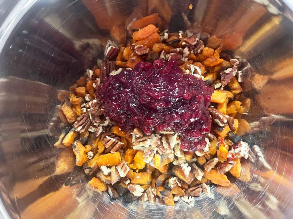 Cranberry sauce added to pecans and sweet potatoes in a mixing bowl.
