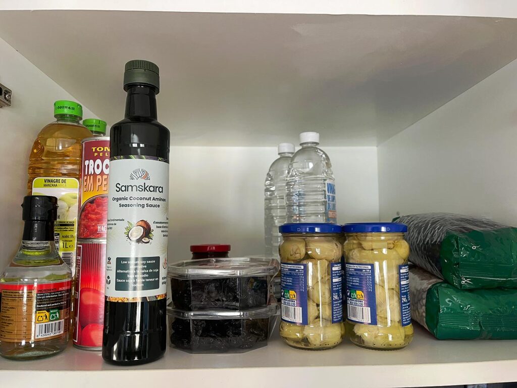 A small cabinet filled with coconut aminos, canned tomatoes, artichoke hearts, vinegars, and dried fruit.