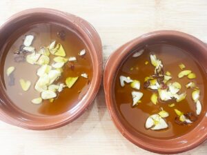 Two Cazuelas filled partway with oil, sliced garlic cloves, and shopped red chilis.
