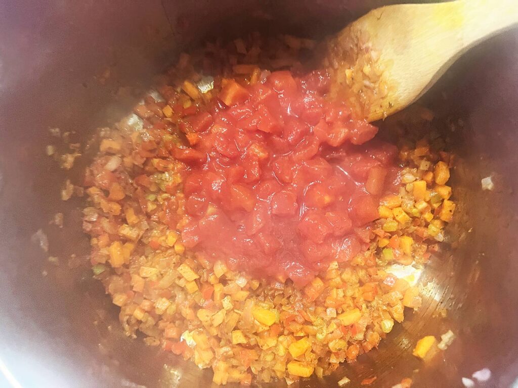 Diced tomatoes added to veggies and spices in a soup pot.