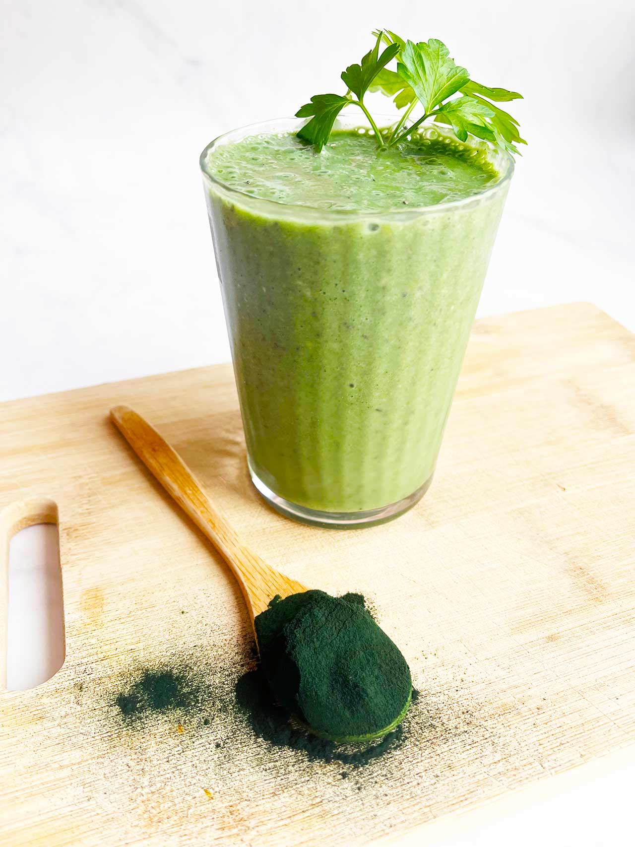 A wood spoon full of Spirulina powder sits in front of a glass of Spirulina Smoothie on a wood cutting board.