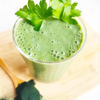 A garnished glass of Spirulina Smoothie holds some greens next to a spoon of spirulina powder.