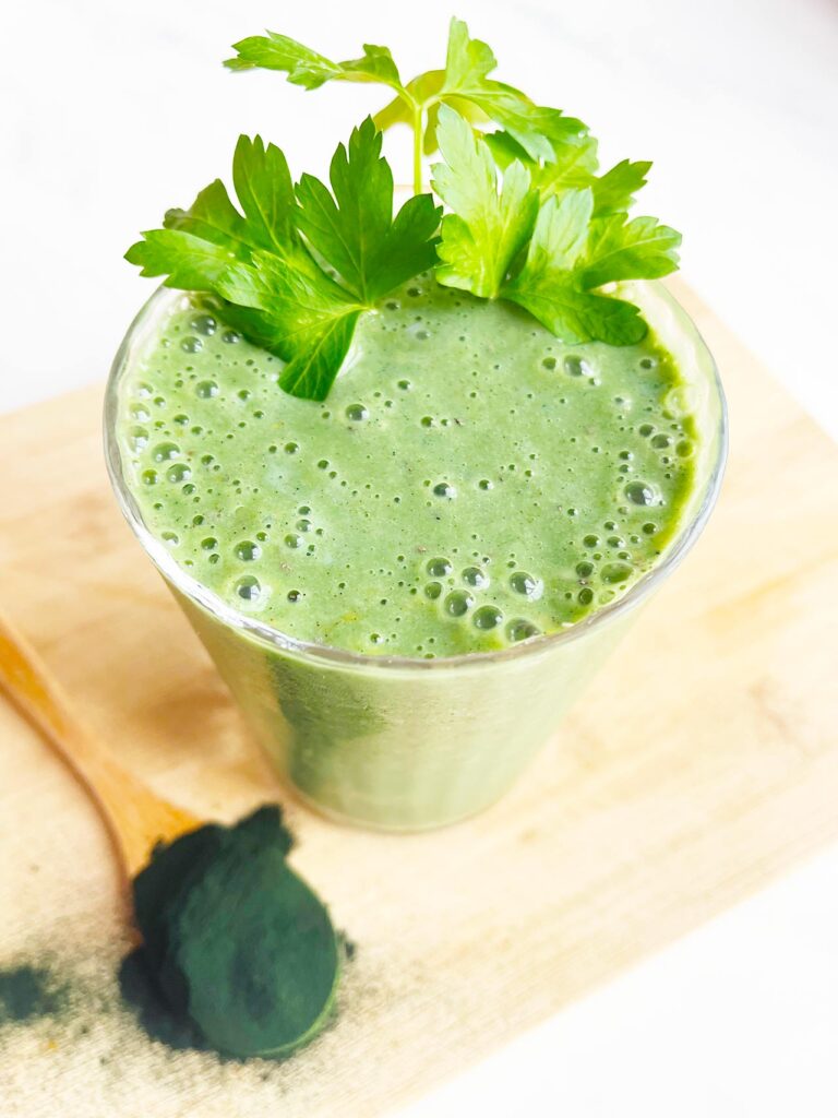 A garnished glass of Spirulina Smoothie holds some greens next to a spoon of spirulina powder.