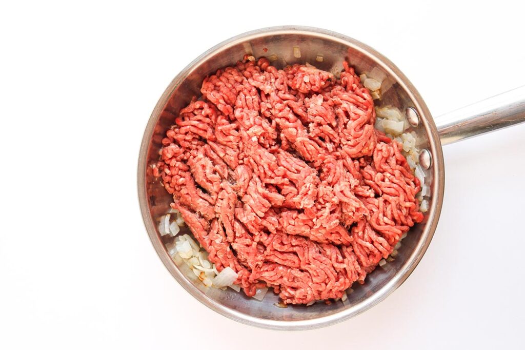 Adding raw, ground beef to saut&eacute;d onions in a skillet.