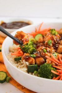 Front, closeup focus. Black chopsticks pick up a pice of teriyaki chicken from a teriyaki chicken bowl.