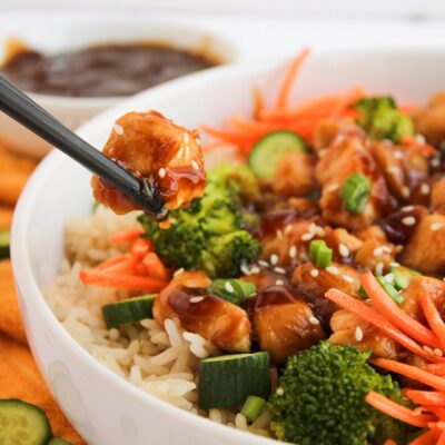 Front, closeup focus. Black chopsticks pick up a pice of teriyaki chicken from a teriyaki chicken bowl.